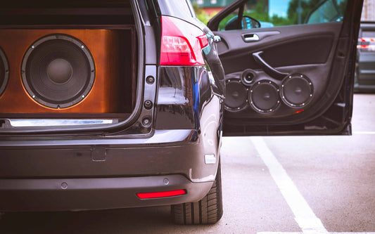 Black car with trunk and rear door open, revealing custom subwoofers and door-mounted speakers in a high-end audio setup