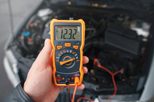 person holding a multimeter reading 12.32 volts near a car engine
