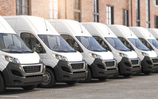 Row of identical white cargo vans parked beside a brick building, suggesting a commercial fleet in an urban setting.