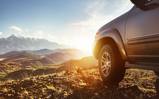 Front of a dark SUV parked on rocky terrain at sunrise or sunset, with scenic mountain ranges in the background.